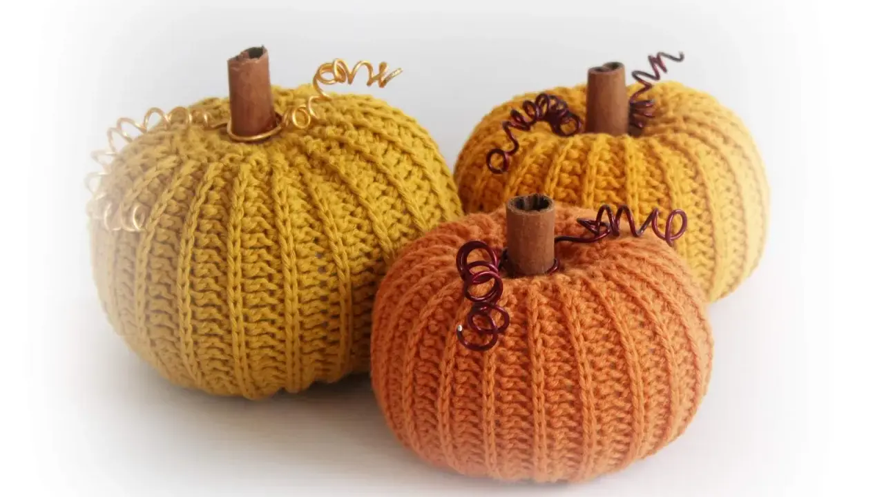 Tre virkade pumpor i höstfärger, en orange och två senapsgula, med kanelstänger som stjälkar och metalltråd som dekoration.