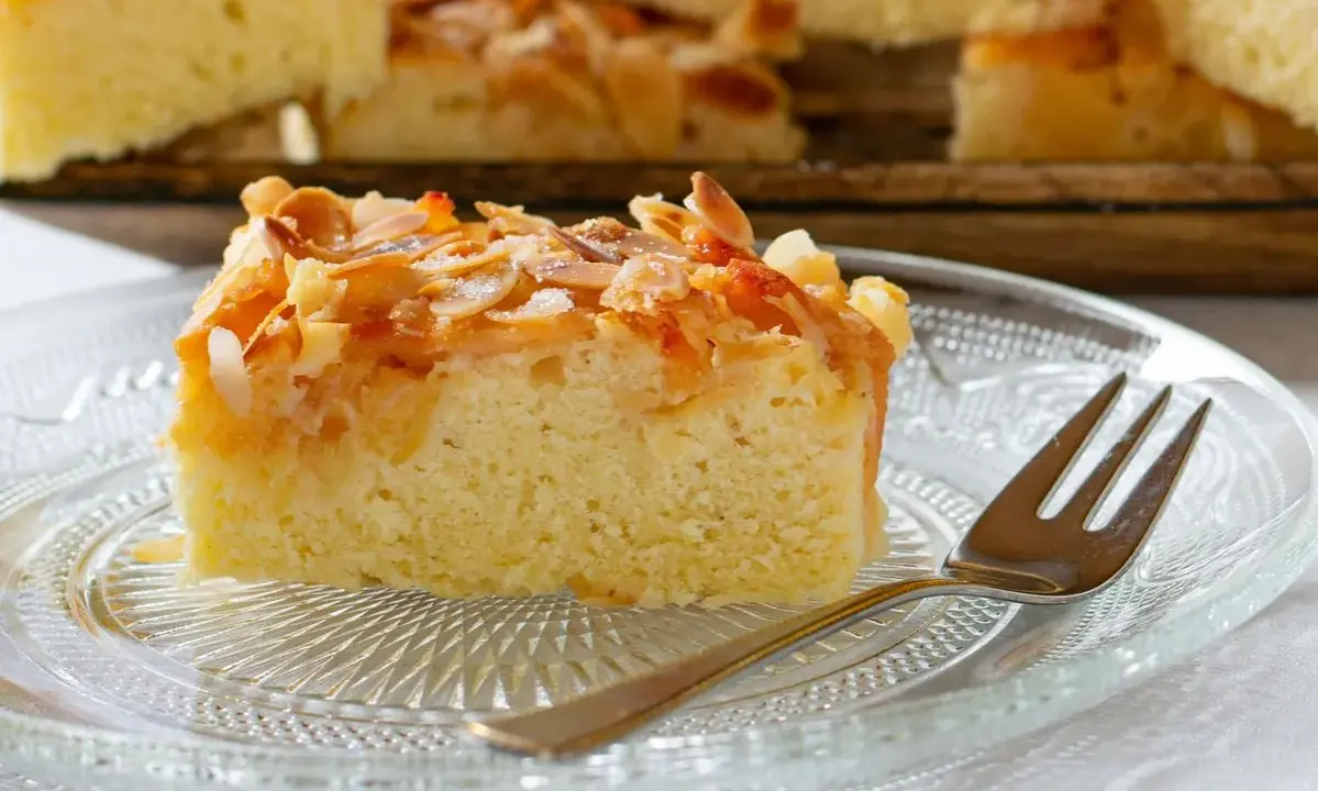 Ein Stück saftiger **Mandelkuchen vom Blech mit gemahlenen Mandeln** und Zucker bestreut, auf einem Glas-Teller mit einer Gabel.