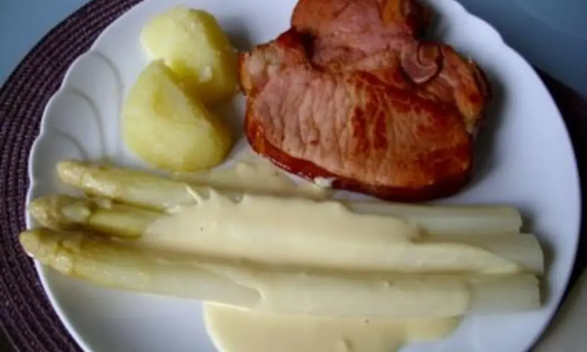 Ein Teller mit gebratenem Schinken, Kartoffeln und Spargel in Sauce Hollandaise. Der Spargel ist in Alufolie gewickelt.