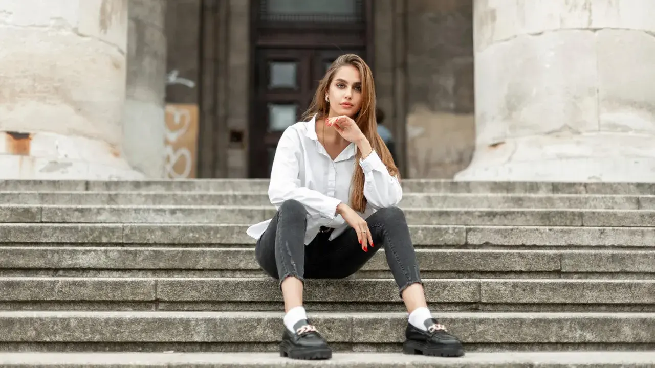 Eleganckie mokasyny to idealne buty do szarych spodni. Kobieta w białej koszuli i czarnych jeansach siedzi na schodach.