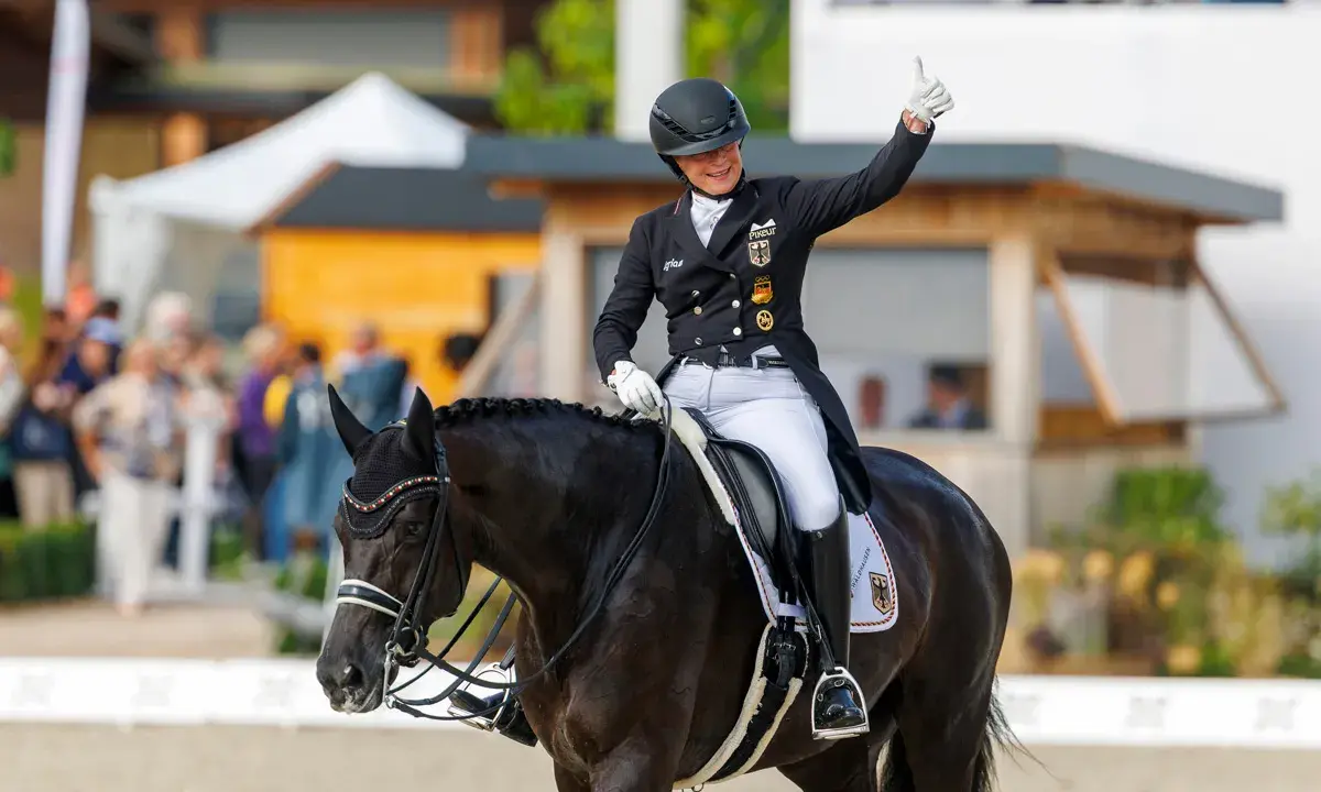 Reiterin gibt Daumen hoch nach erfolgreichem Grand Prix Reiten. Dunkles Pferd mit Turnieroutfit und Helm.