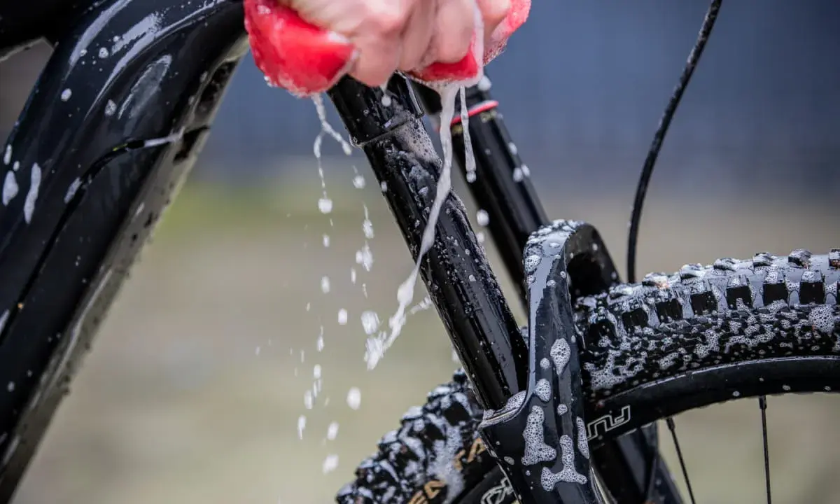 Mit Hausmitteln das Fahrrad sauber machen: Eine Hand mit rotem Schwamm reinigt den Rahmen und die Federgabel, Schaum und Wasser spritzen.
