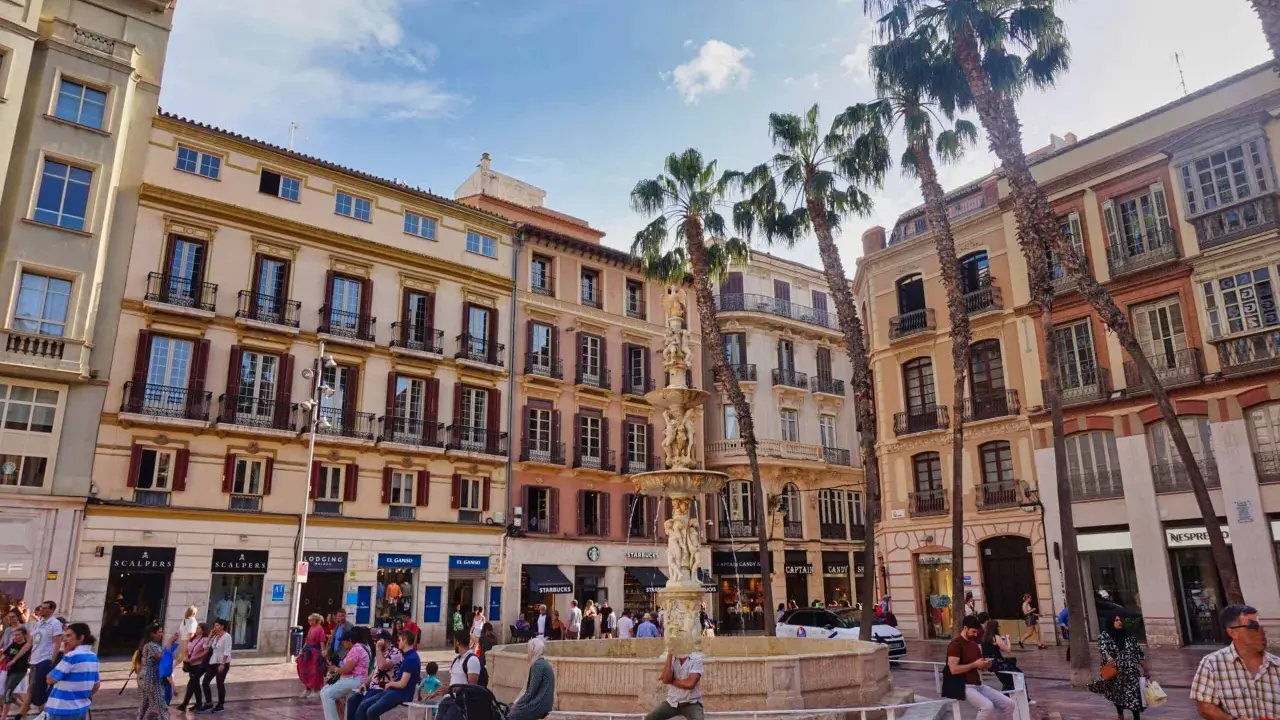 Sonniger Platz in der **Malaga Altstadt** mit einem Brunnen, Palmen und bunten Gebäuden. Menschen flanieren und genießen die Atmosphäre.