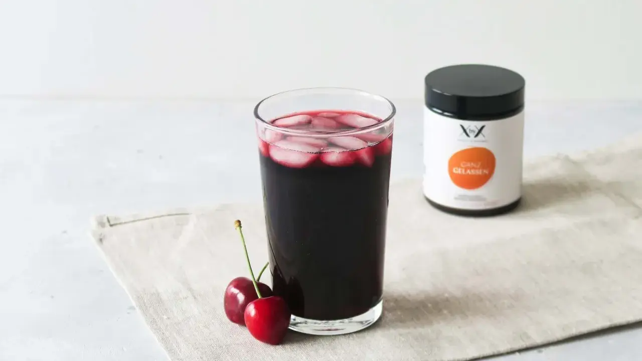 Ein Glas dunkler Sauerkirschsaft mit Eiswürfeln und zwei Kirschen daneben. Eine Dose mit der Aufschrift "Ganz Gelassen" steht im Hintergrund. Die positive sauerkirschsaft wirkung wird hier visuell dargestellt.