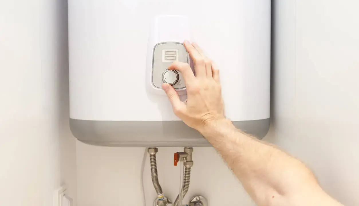 Mano ajustando el control de temperatura de un termo eléctrico. La instalación se ve limpia y moderna.
