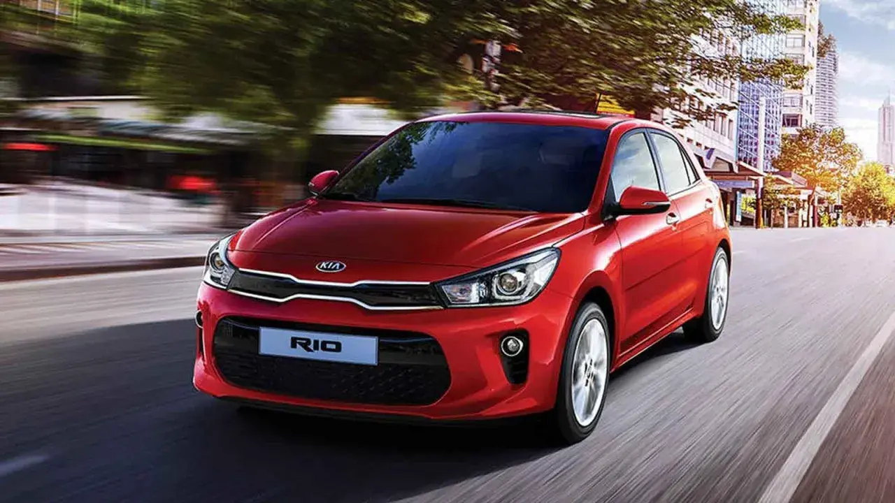Un hatchback rojo Kia Rio se desplaza por una calle urbana, con edificios y árboles borrosos por el movimiento.