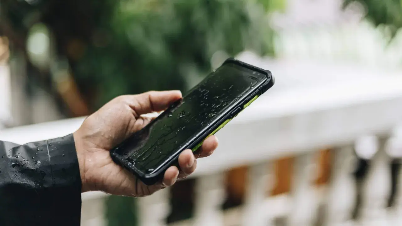 Dłoń trzyma mokry smartfon ip67 na tle zieleni. Krople deszczu spływają po ekranie.