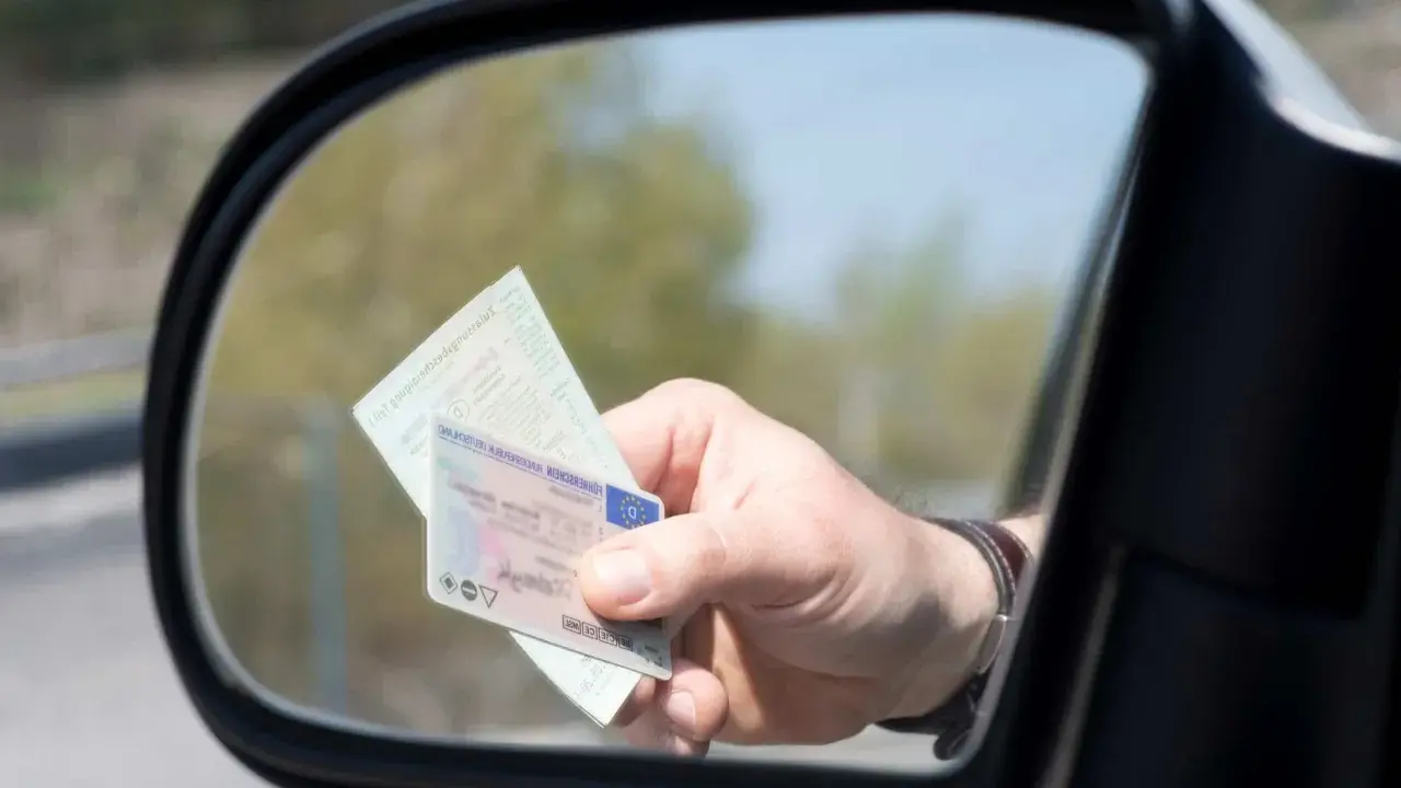 Mano sujetando un carnet de conducir y un documento de identidad reflejados en el espejo retrovisor de un coche.