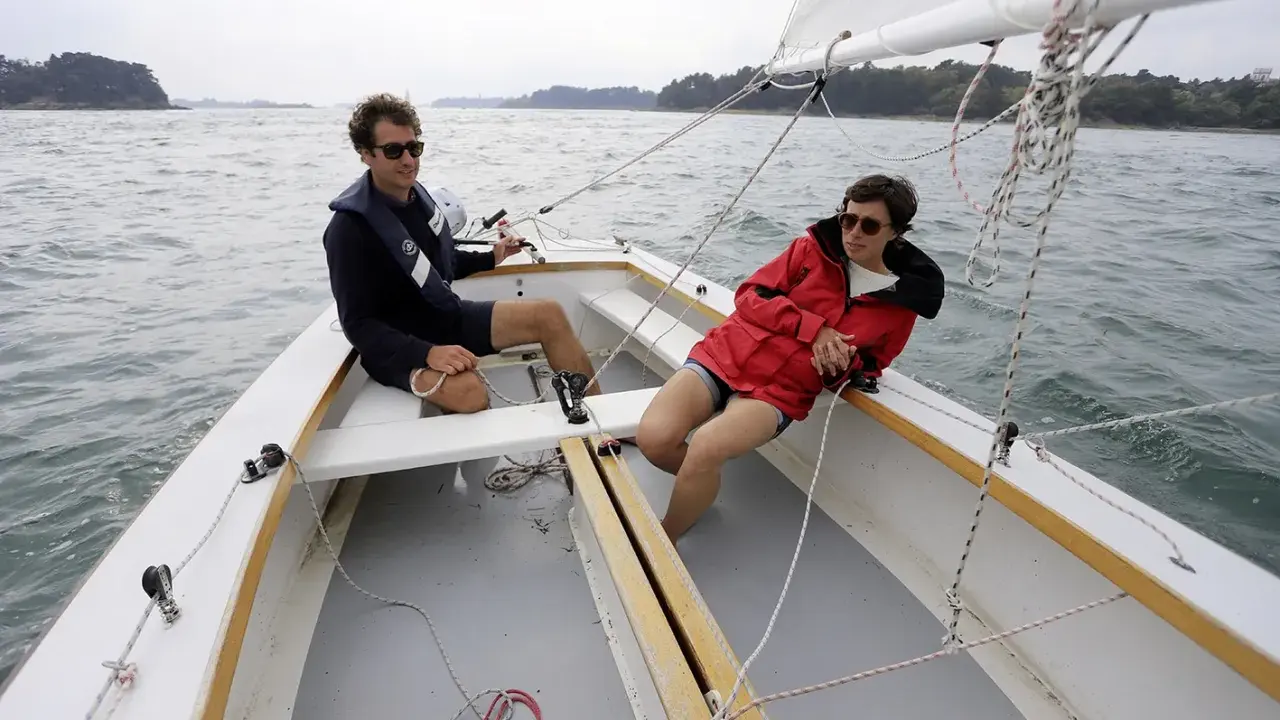 Deux personnes profitent d'une journée ensoleillée sur un voilier dayboat, naviguant sur une mer calme.