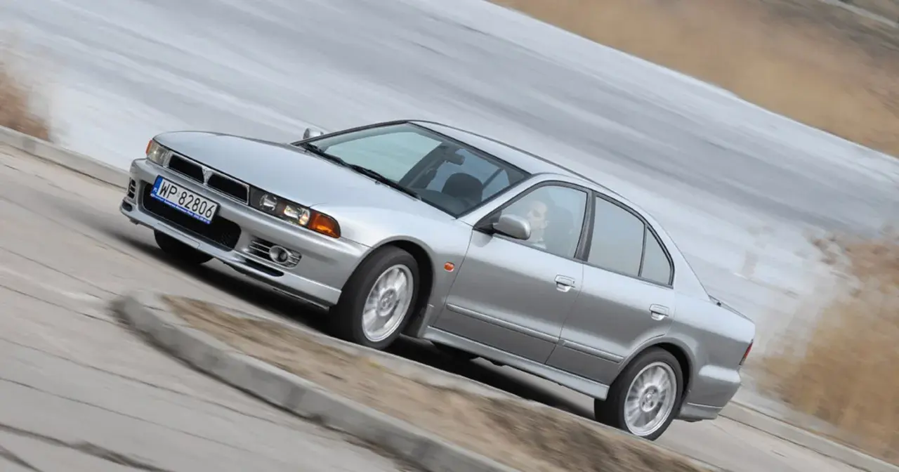 Srebrny Mitsubishi Galant z gracją pokonuje zakręt. Kierowca w okularach pewnie prowadzi auto.