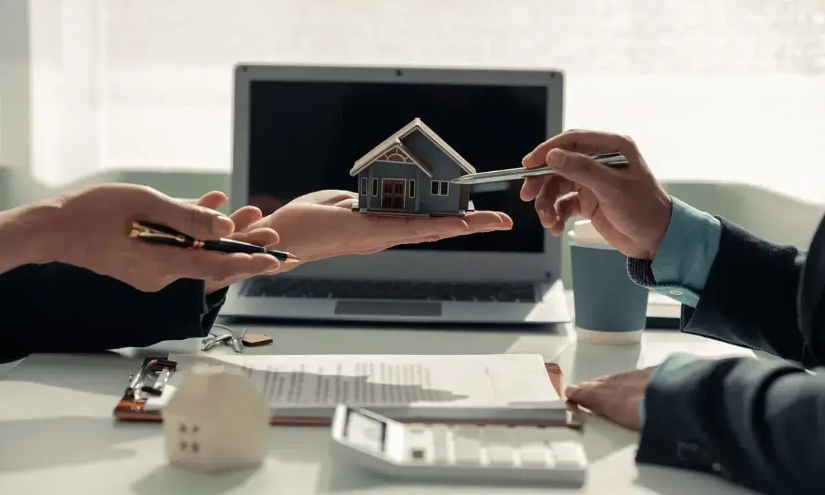 Deux mains présentent une maquette de maison, symbolisant comment devenir investisseur immobilier. Un contrat et un ordinateur sont visibles.