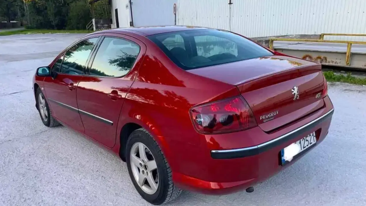 Błyszczący czerwony Peugeot 407 2.0 HDI z przyciemnianymi szybami stoi na żwirowym podjeździe.