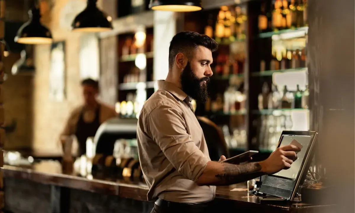 Hombre con barba gestionando un bar, usando una tablet y una pantalla táctil para procesar pagos.