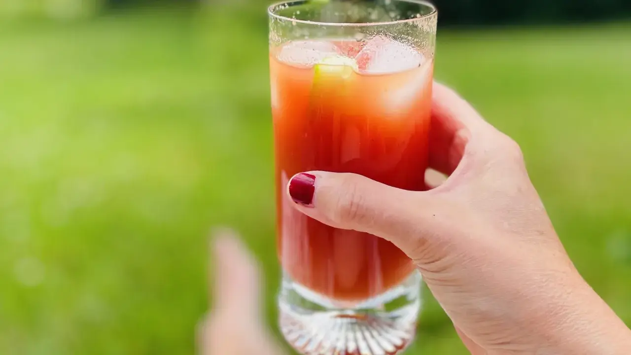 A hand with red nail polish holds a tall glass of Bloody Mary with ice and a cucumber slice, outdoors.