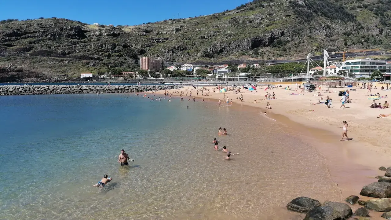 Sonniger Strand in Machico, Madeira, mit vielen Menschen, die im klaren Wasser planschen und sich am Sandstrand sonnen.