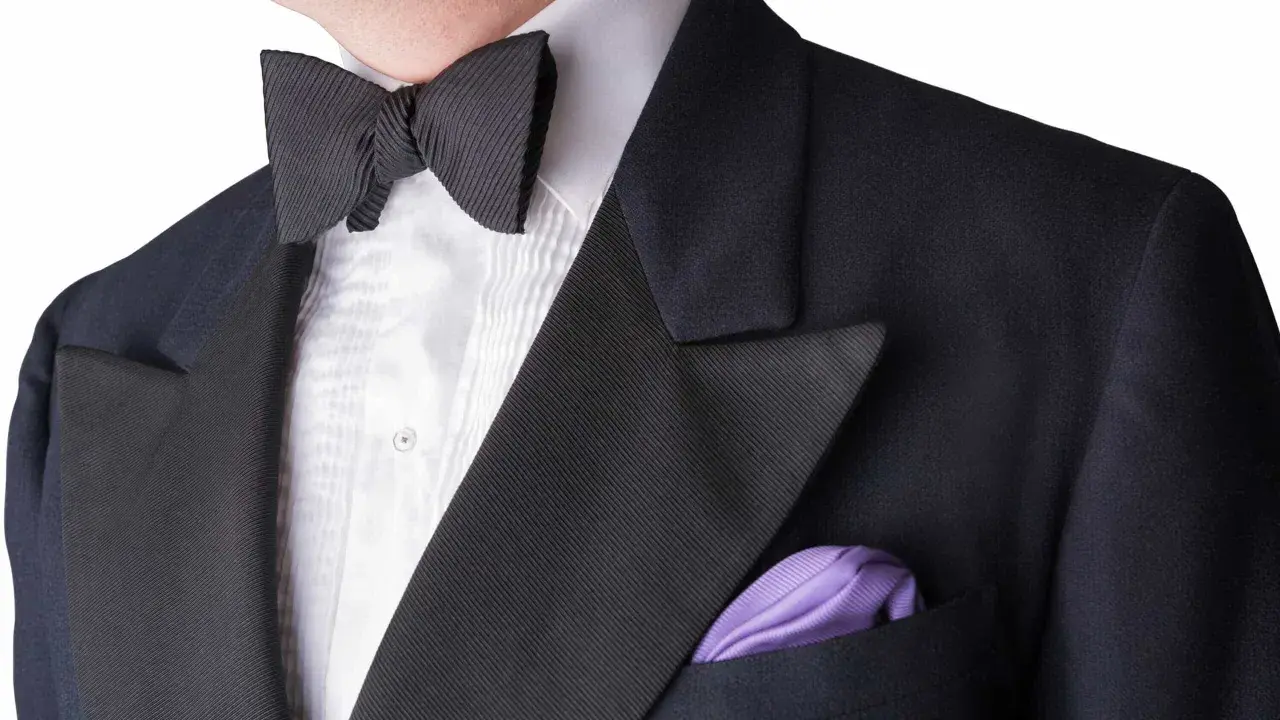 A man wears a black tuxedo with a pleated white shirt and a black bow tie. A purple pocket square adds a pop of color. This outfit answers the question: can you wear a tie with a tux? (No, a bow tie is traditional.)