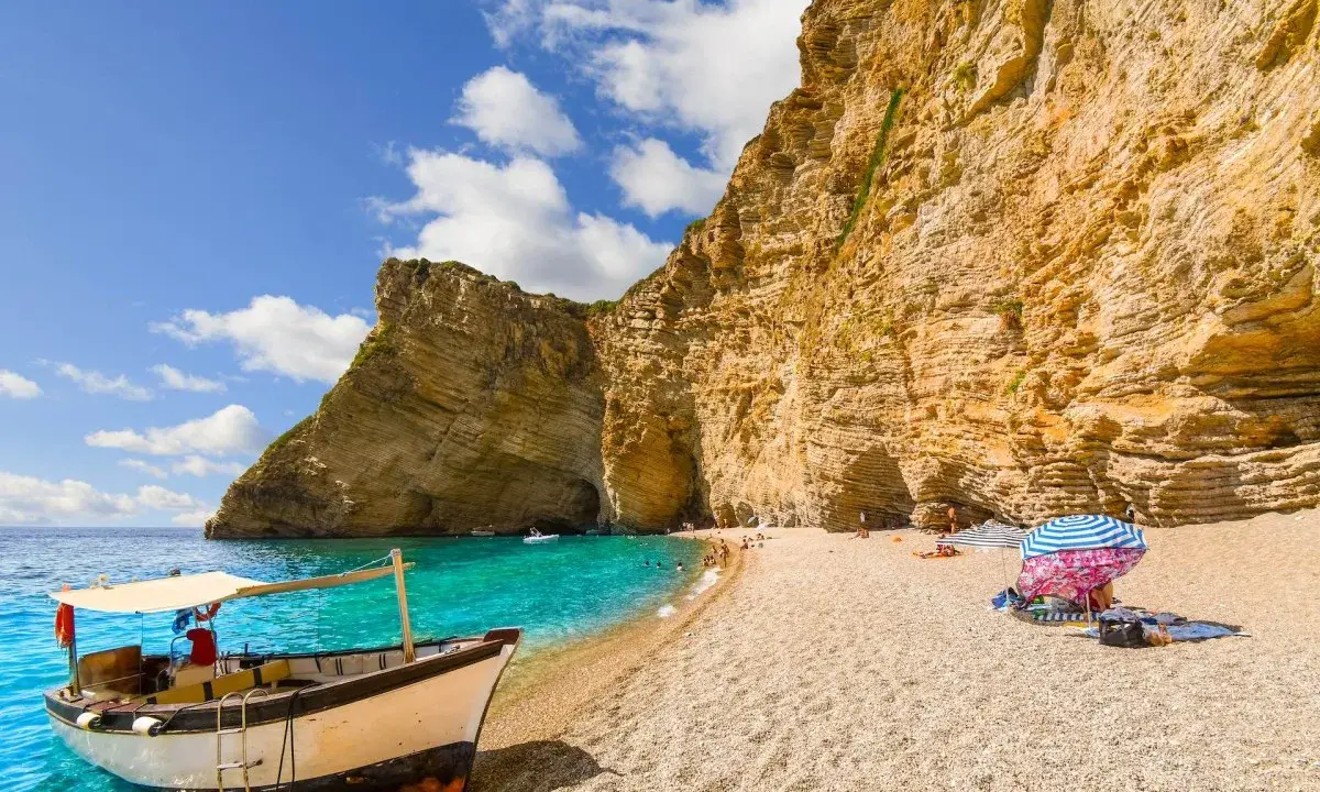 Ein Boot liegt am Korfu Strand vor einer beeindruckenden Felswand. Türkises Wasser und ein sonniger Himmel laden zum Verweilen ein.