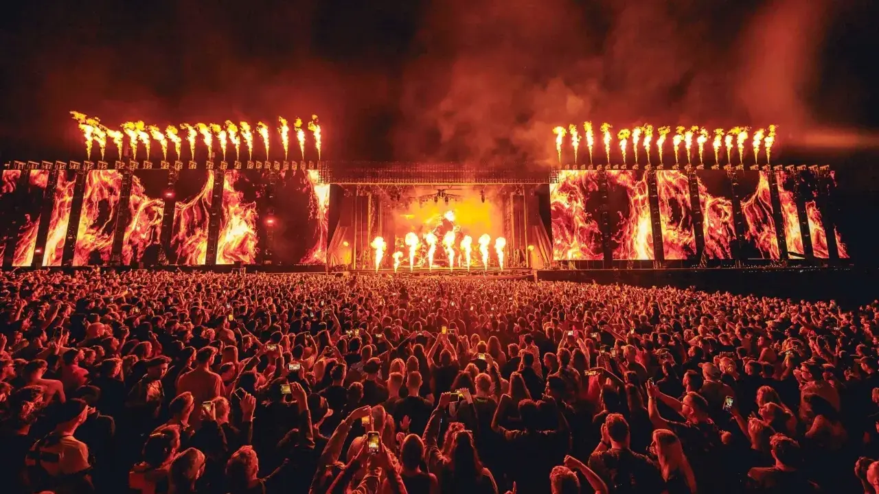Un'immensa folla partecipa a un festival tekno, illuminata da fiammate spettacolari e schermi con immagini di fuoco.