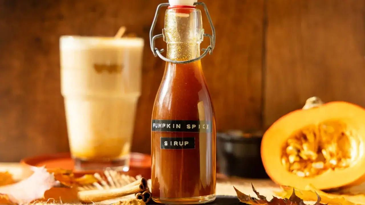 Flasche mit goldbraunem Pumpkin Spice Sirup, daneben ein Latte Macchiato, Kürbishälften und Herbstlaub.