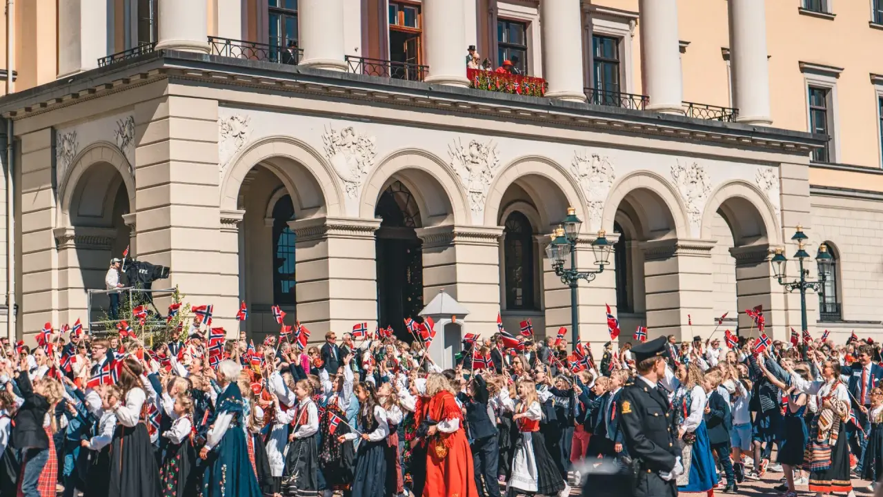 Tłum ludzi, w tym dzieci, świętuje, machając norweskimi flagami. Wiele osób nosi tradycyjny strój norweski, ozdobiony haftami i kolorowymi wzorami.