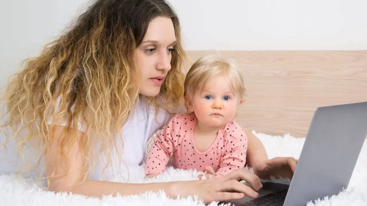 Kobieta z dzieckiem na rękach pracuje na laptopie, zastanawiając się, czy na urlopie wychowawczym można pracować.