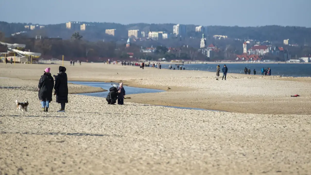 Spacerem po piaszczystej plaży w Jelitkowie, z widokiem na morze i miasto w tle. Ludzie spacerują, bawią się z psem.