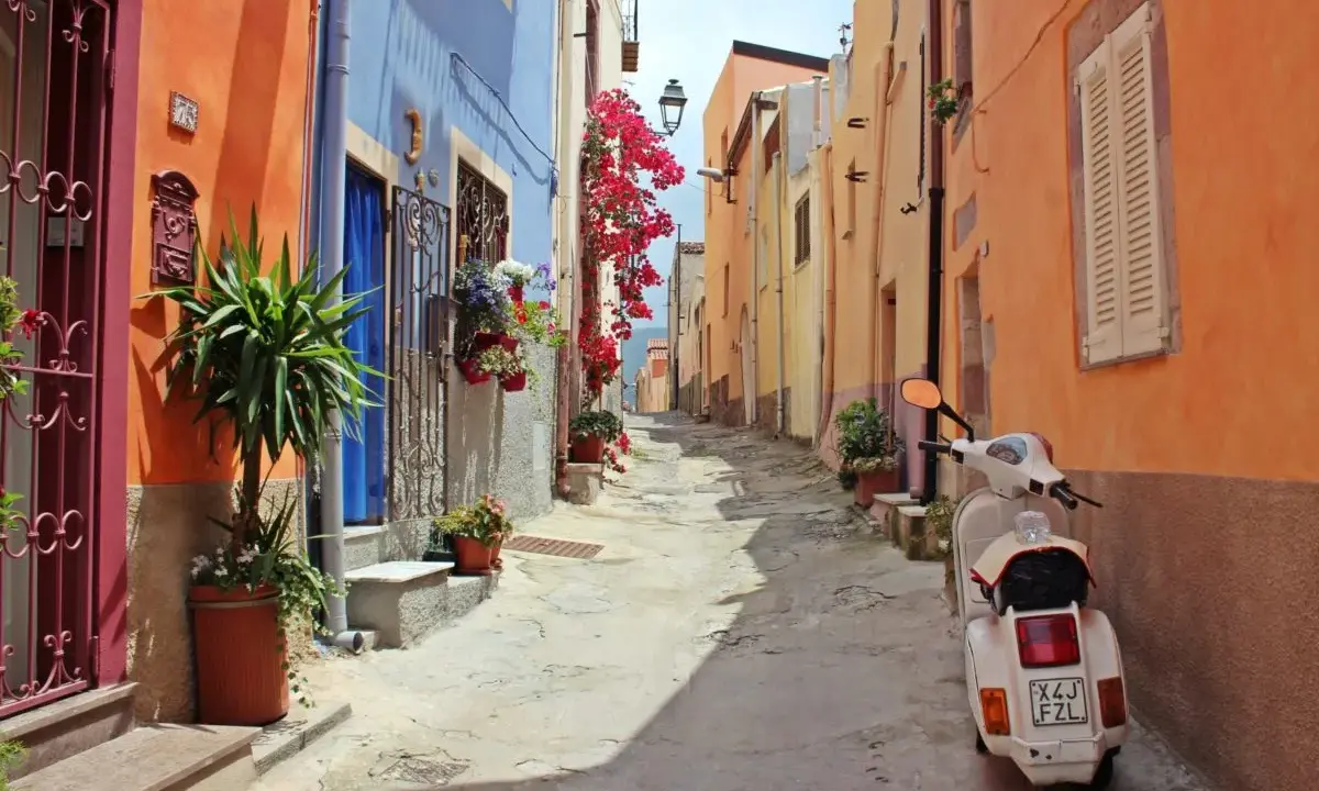 Un vicolo colorato con case arancioni e blu, fiori rossi e una Vespa bianca. Perfetto per le tue idee weekend nord Italia.