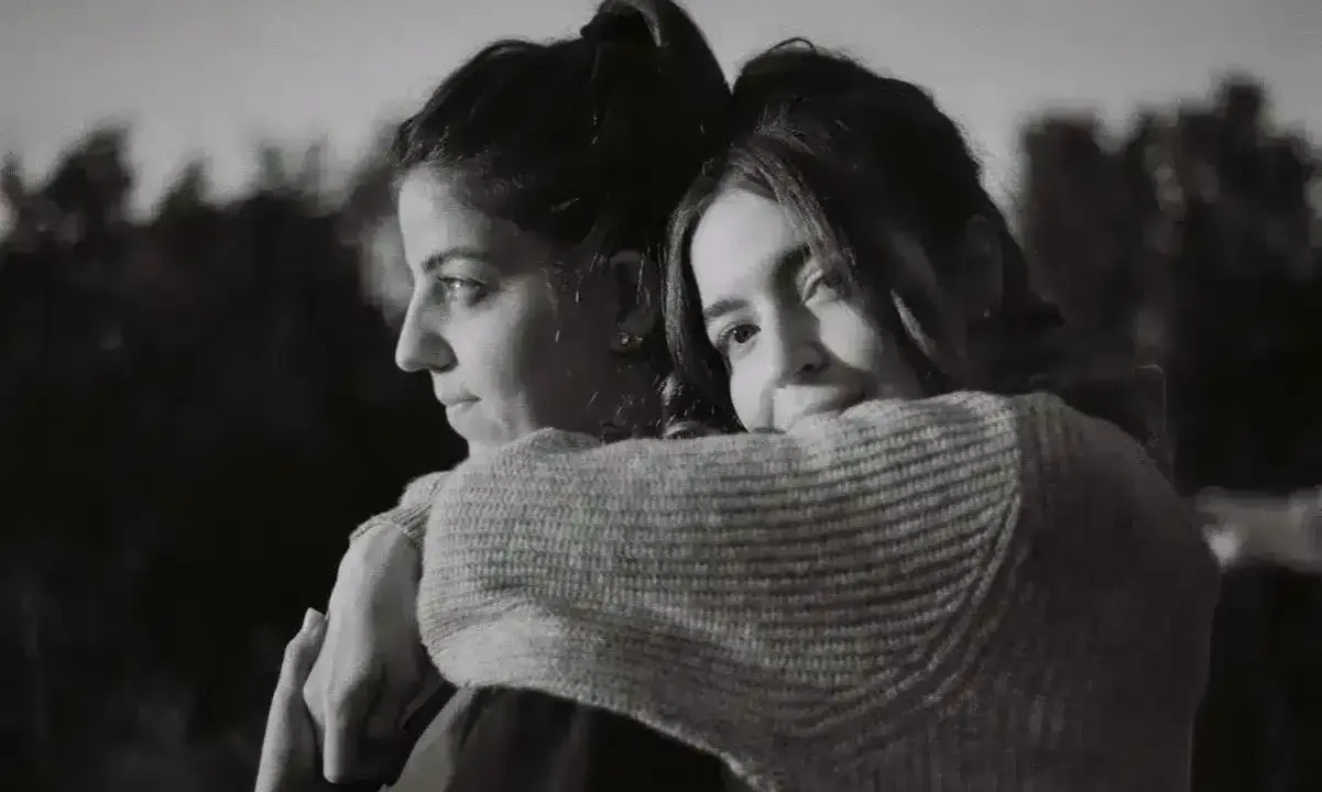 Dos mujeres se abrazan, una mirando al horizonte con **miedo al abandono**, la otra la abraza con ternura.