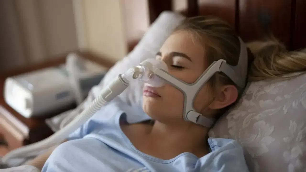 Kobieta śpi w masce CPAP, która zapewnia wspomaganie oddychania w domu. Urządzenie pomaga jej spokojnie przesypiać noce.