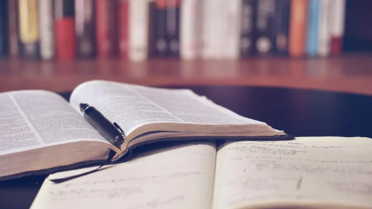 Otwarta księga i notatnik na tle regału z książkami. Idealne do nauki lektury liceum.