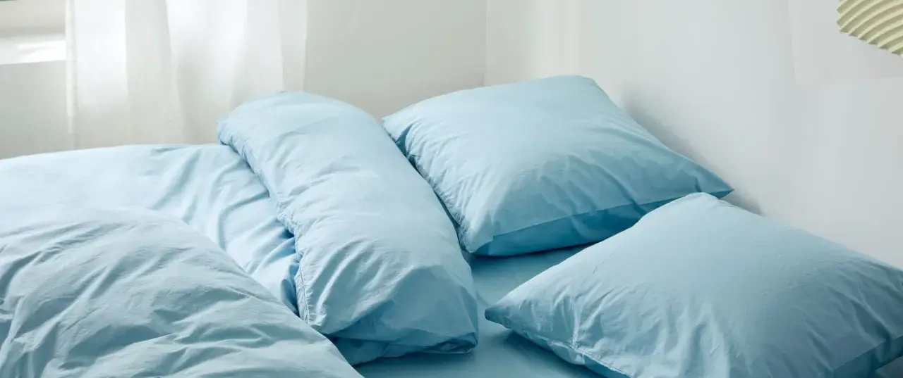 En bädd med ljusblå sängkläder, troligen percale eller satin, med flera kuddar och ett täcke.
