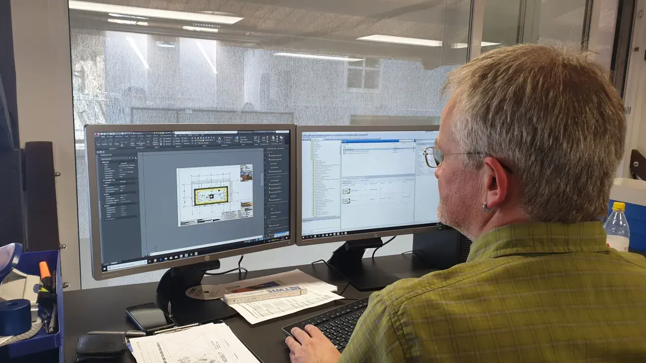 Mann am Computer, der Baupläne nach DIN 4426 erstellt. Auf dem Bildschirm sind CAD-Zeichnungen und eine Liste von Bauteilen zu sehen.