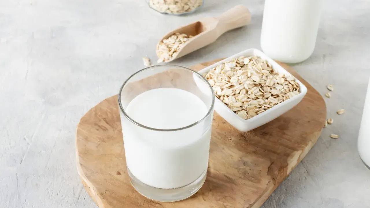 Ein Glas cremiger Hafermilch steht auf einem Holzbrett, daneben Schüsseln mit Haferflocken. Hafermilch gesund und lecker!