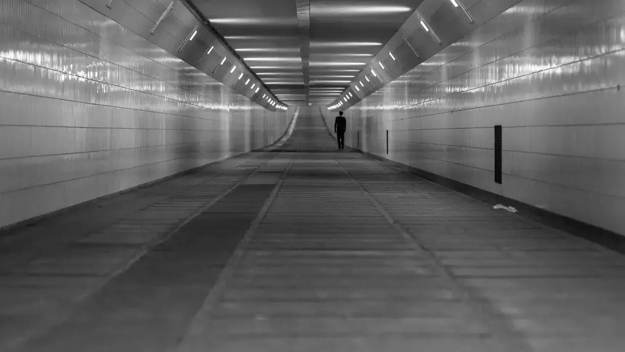Człowiek w garniturze idzie przez długi, pusty tunel. Światła na ścianach tworzą wrażenie nieskończoności, jakby wchodził w szarą strefę.