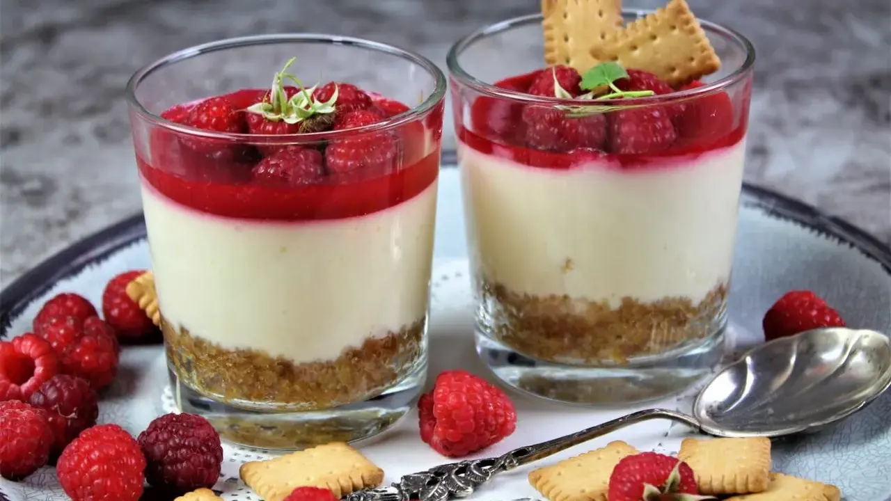 Zwei Gläser mit kalorienarmen Desserts im Glas: cremige Schicht, Himbeersoße und Keksboden. Garniert mit frischen Himbeeren und Keksen.