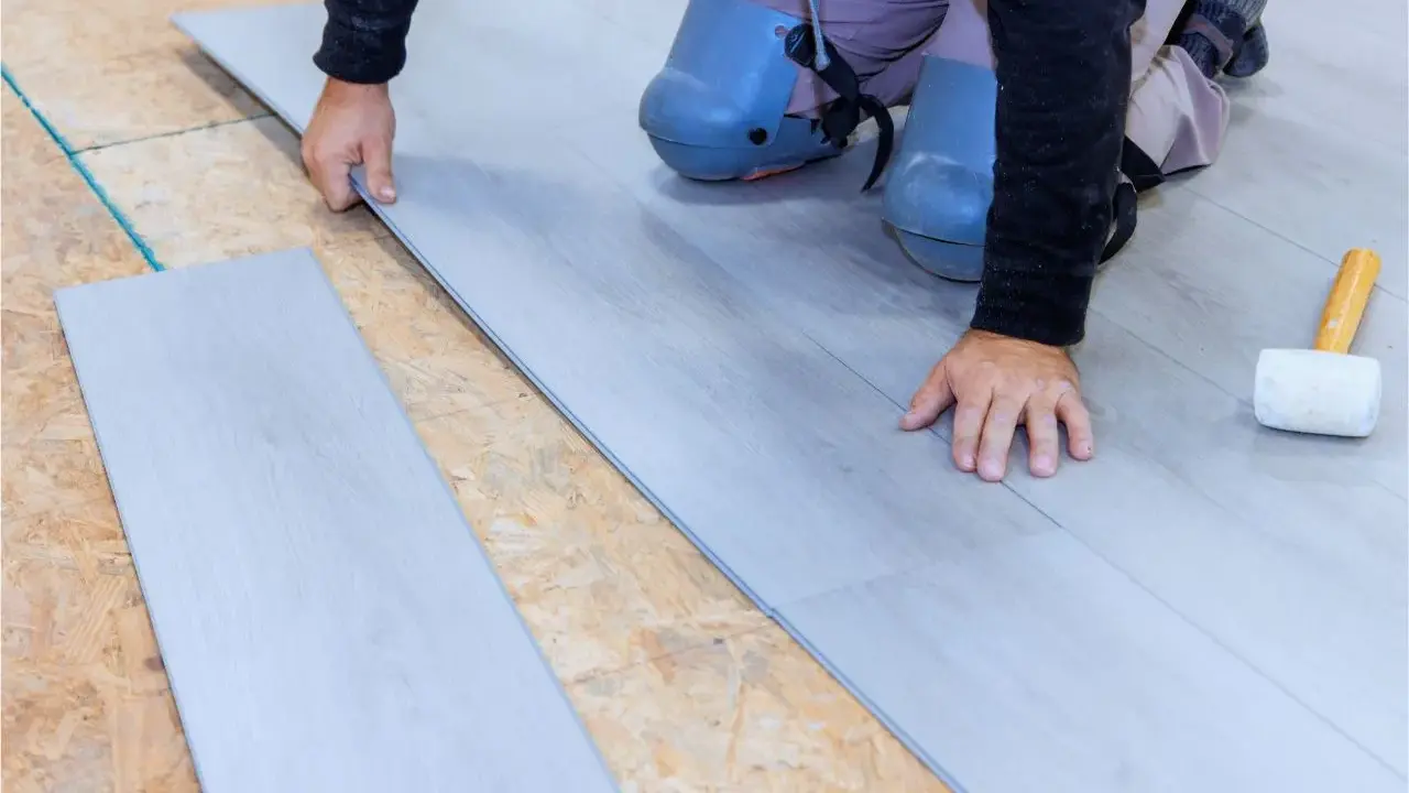 Man con rodilleras coloca cuidadosamente tablones de suelo laminado gris sobre una base de madera.
