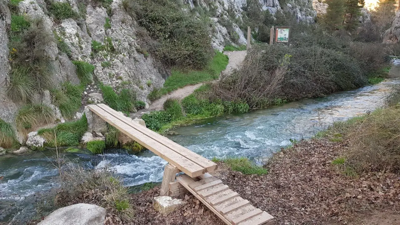 Puente de madera sobre un río caudaloso, parte de la ruta del zarzalar. Senderos y vegetación agreste flanquean el camino.