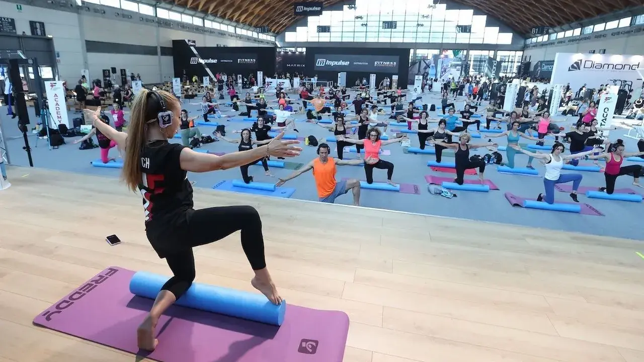 Allenamento di gruppo alla fiera dello sport Rimini, con istruttrice e partecipanti su tappetini.