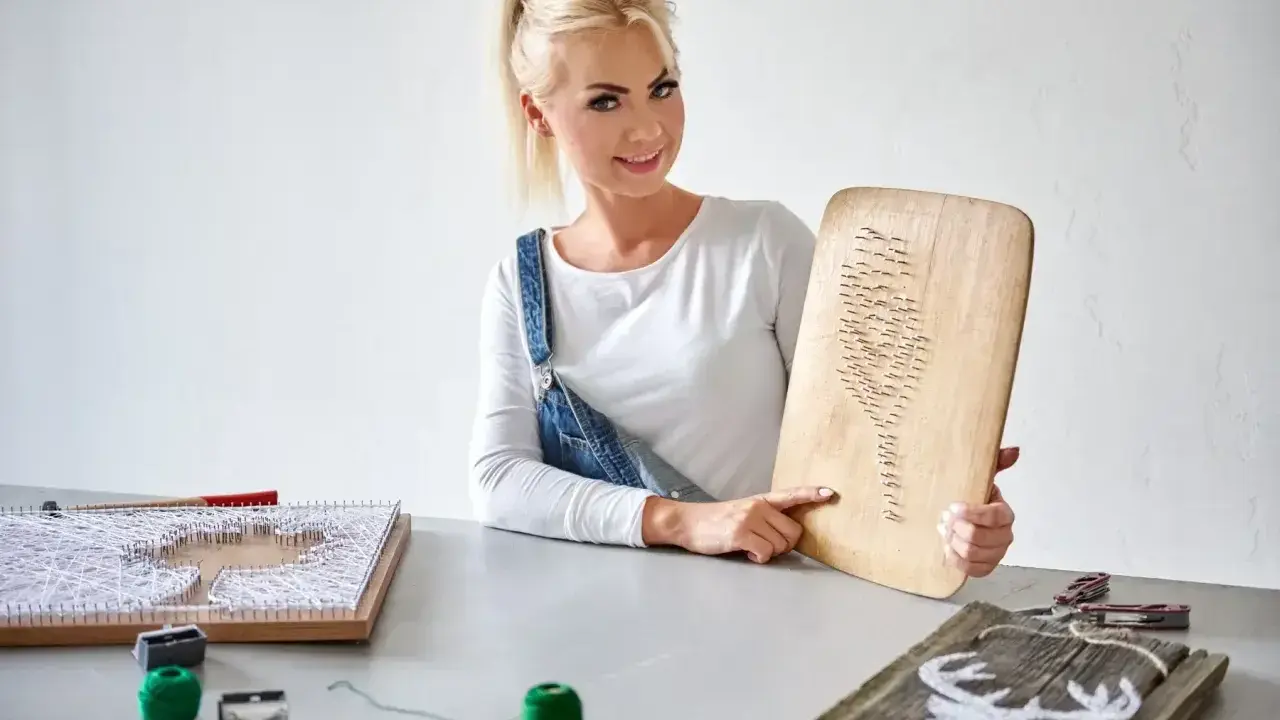 Kobieta prezentuje obraz z nici i gwoździ szablon. Na stole widać inne prace i materiały do tworzenia.