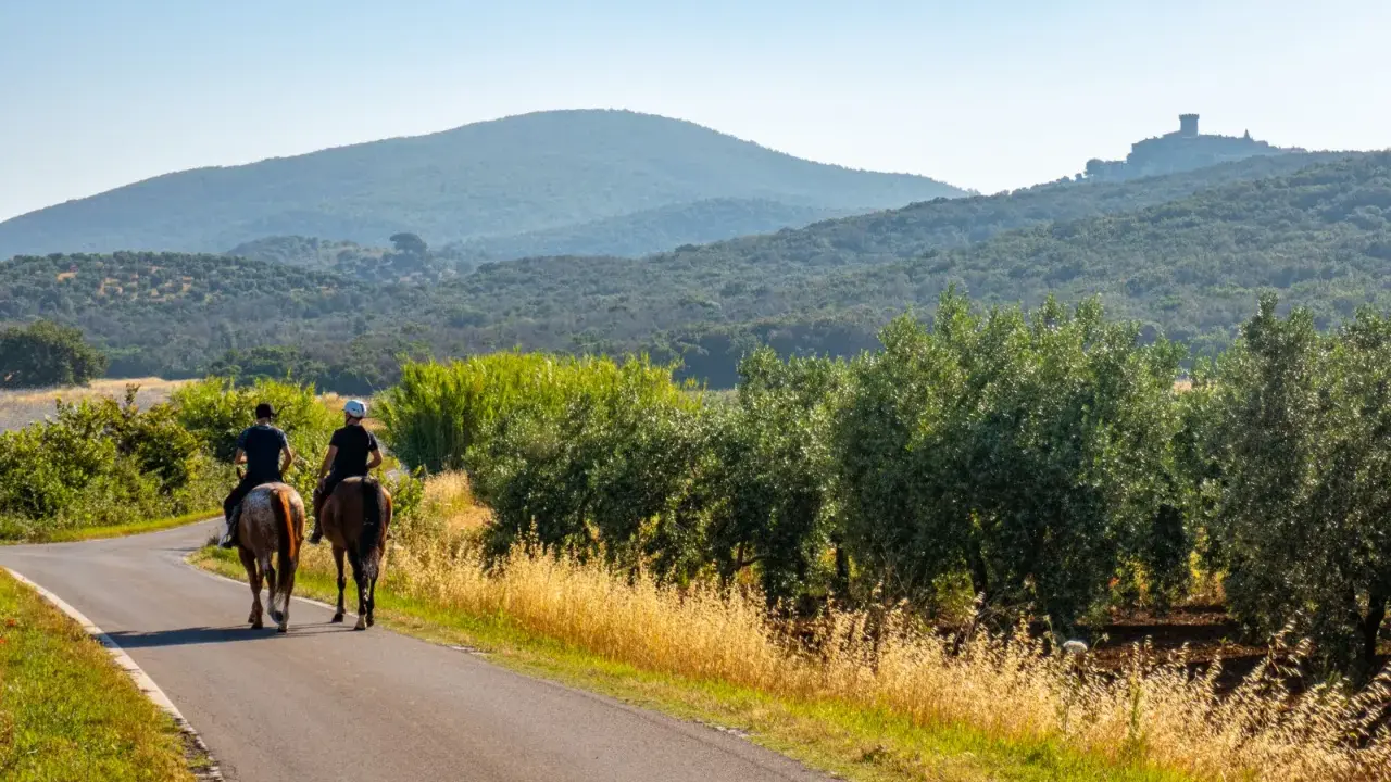 Due cavalieri godono di una **passeggiata a cavallo toscana** tra ulivi e colline, con un castello in lontananza.