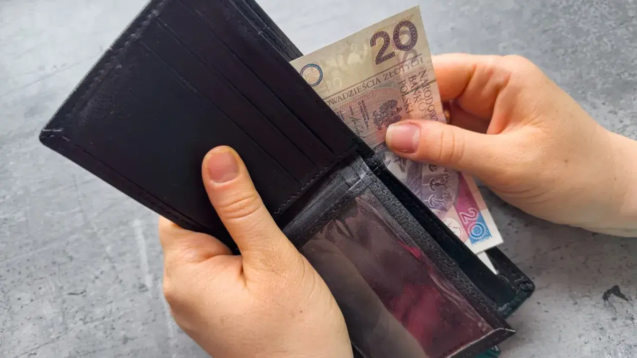Ręce wkładają banknot 20 zł do portfela. Dowiedz się, komu przysługuje zasiłek dla bezrobotnych.