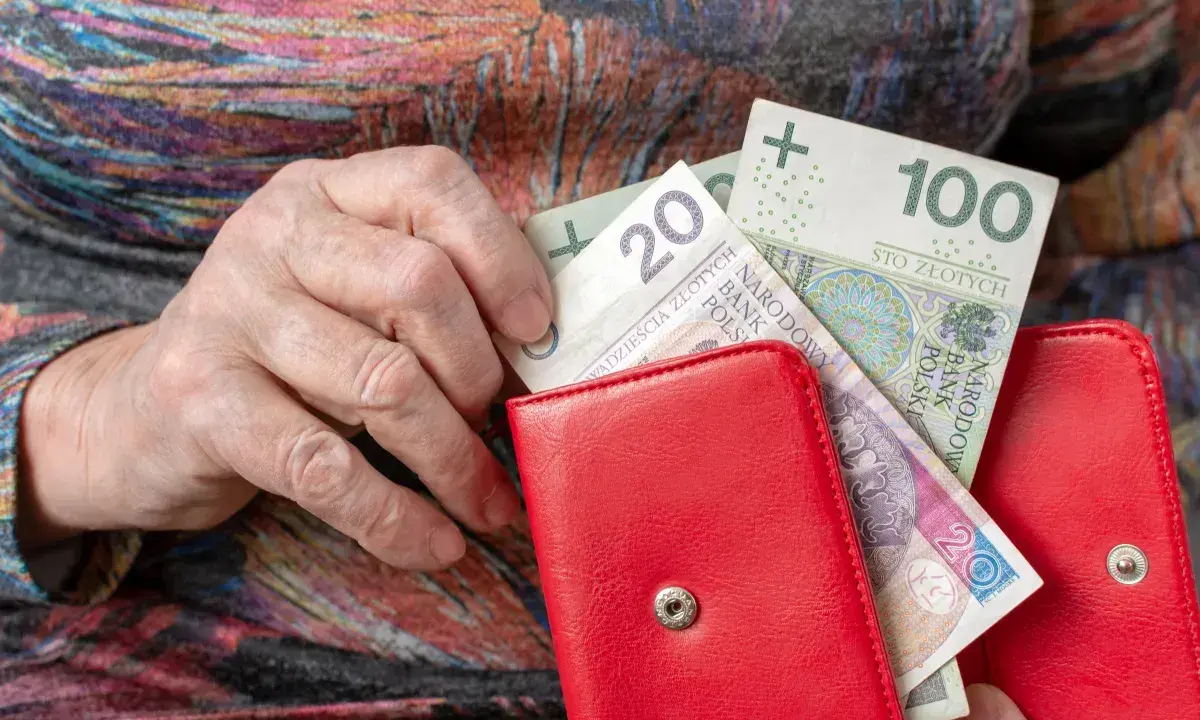 Dłoń starszej osoby wkłada pieniądze do czerwonego portfela. Może to być dodatek mieszkaniowy.