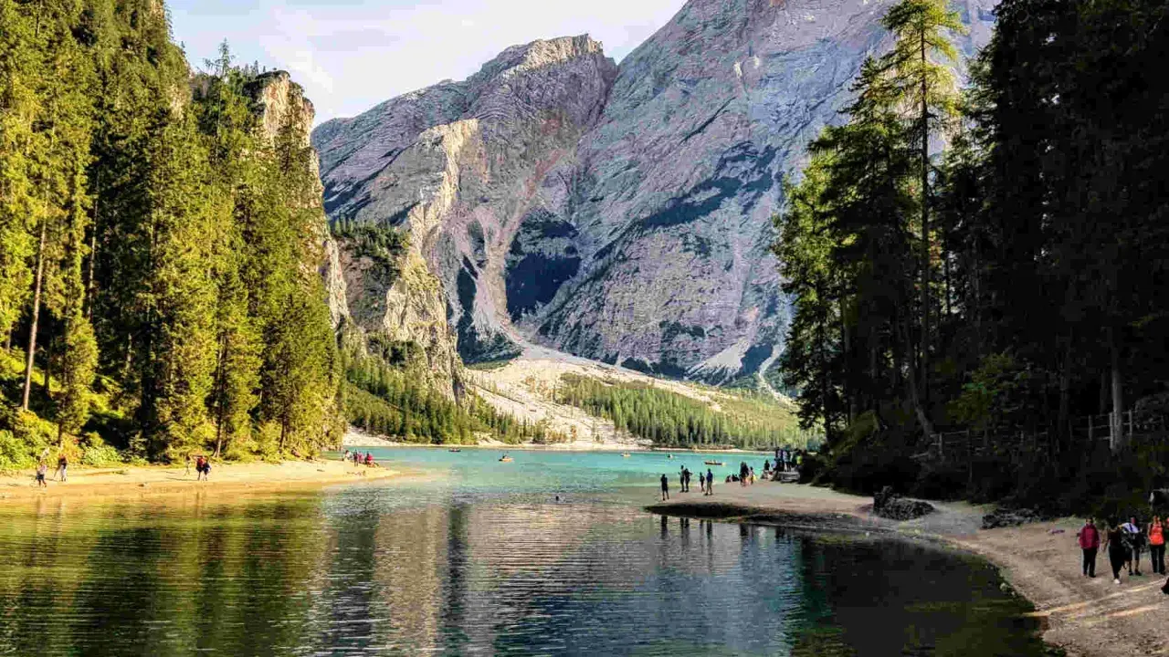 Sentiero da Valdaora al Lago di Braies: acque turchesi, montagne imponenti e pinete rigogliose. Turisti passeggiano sulla riva sabbiosa.