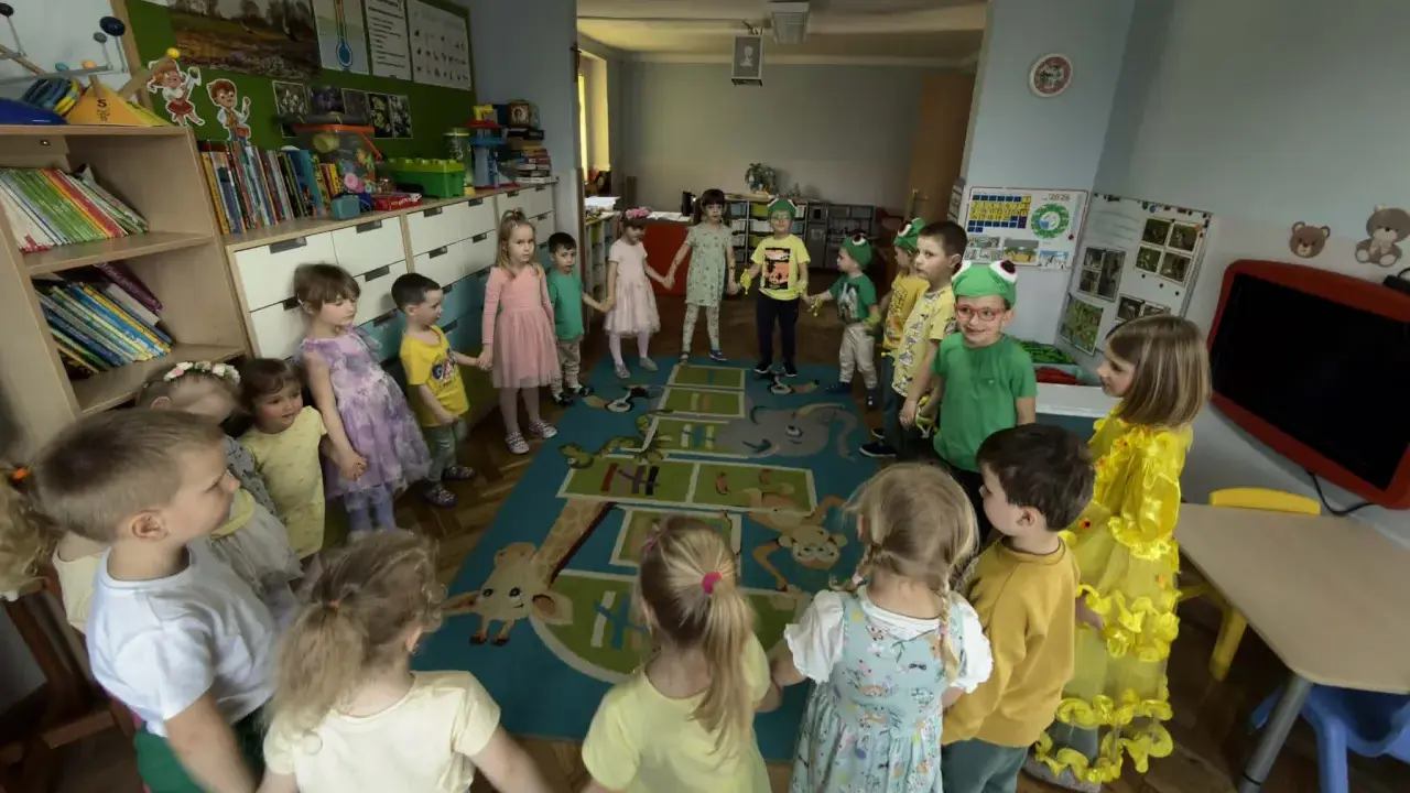 Dzieci trzymają się za ręce, tworząc krąg na dywanie z zabawnymi zwierzątkami. To radosna zabawa w **zespole szkolno przedszkolnym**.