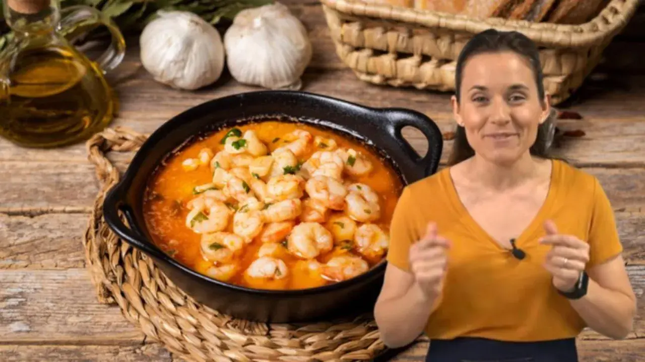 Una mujer sonríe mientras muestra cómo hacer gambas al ajillo, con un plato humeante y los ingredientes listos.