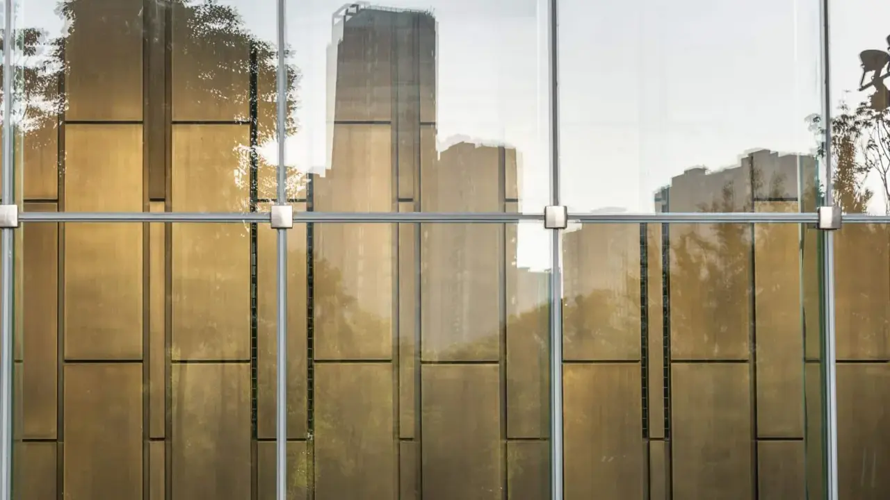 Reflejos dorados de árboles y edificios en un **cristal blindado** moderno, con estructura metálica visible.