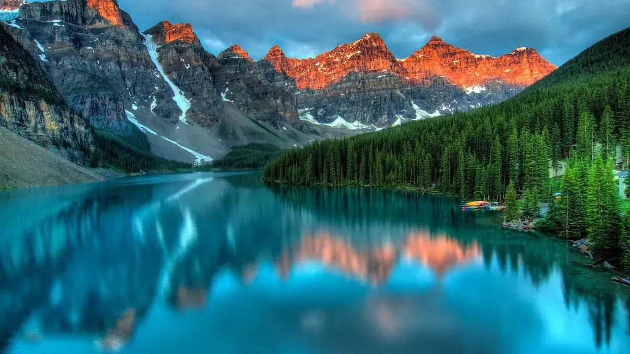 Turkusowe jezioro odbija ośnieżone szczyty gór o zachodzie słońca. W tle gęsty las iglasty. Park narodowy Banff zachwyca.
