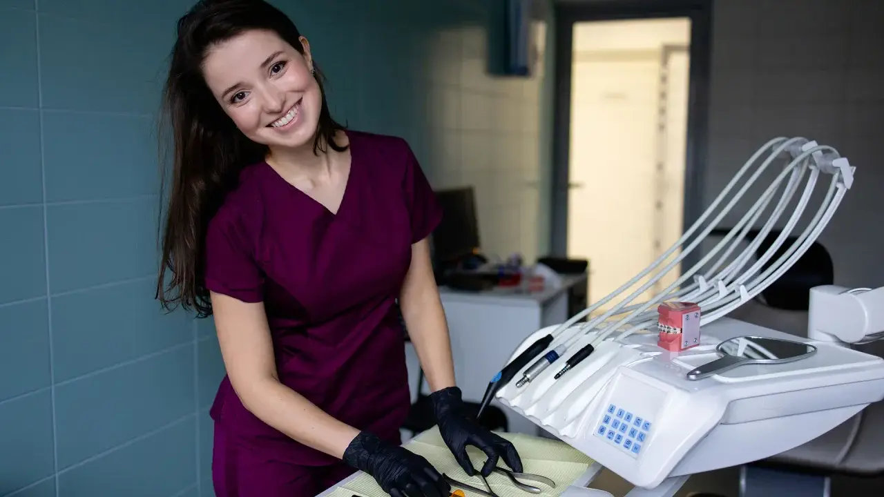 Uśmiechnięta asystentka stomatologiczna w bordowym uniformie przygotowuje narzędzia na tacce obok fotela dentystycznego.