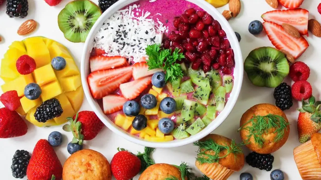Kolorowa miska smoothie z owocami, jagodami, granatem i wiórkami kokosowymi. Obok mango, truskawki, kiwi, maliny i muffinki. Idealne owoce na noc.