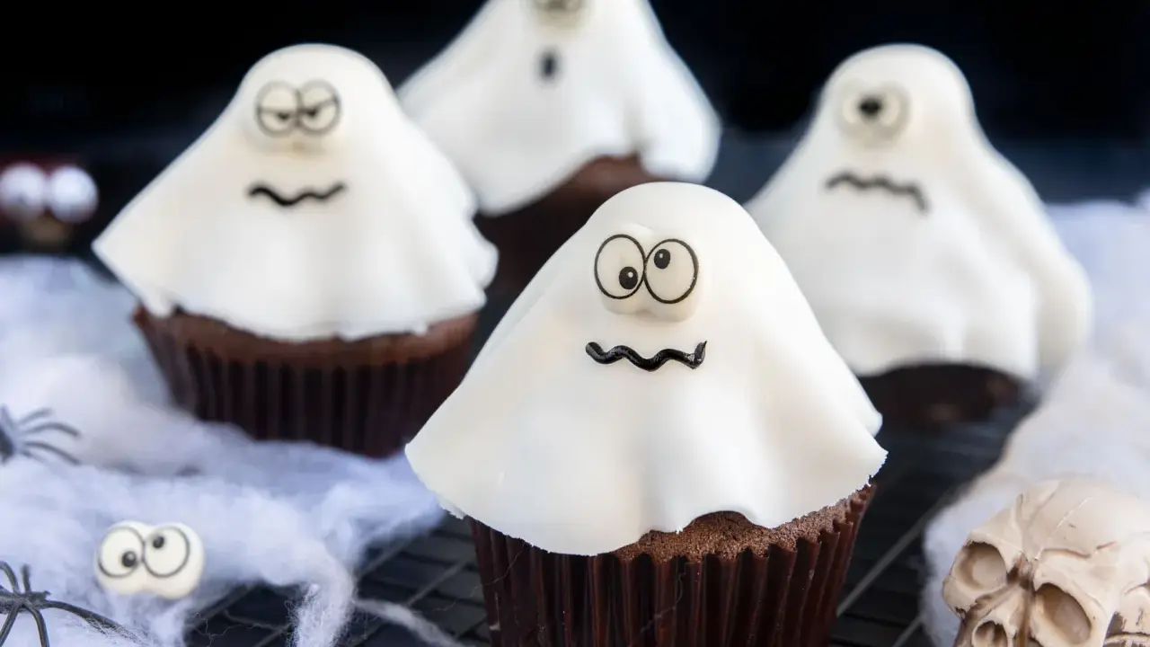 Vier schokoladige gespenster muffins mit weißen Geisterhauben und lustigen Gesichtern, dekoriert mit Spinnweben und einem Totenkopf.